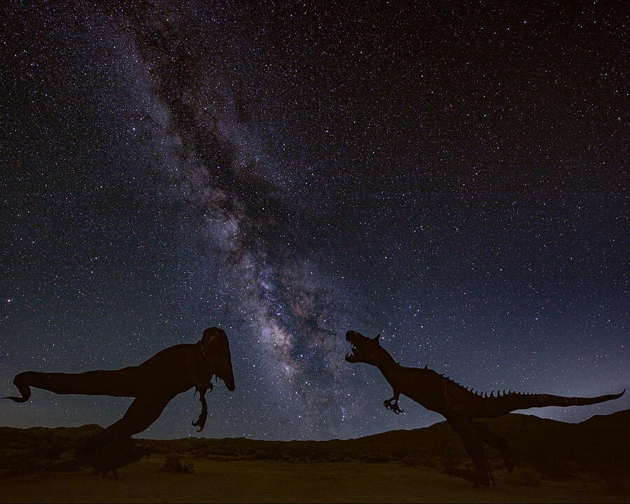 Two dinosaur silhouettes face each other beneath the Milky Way in a desert nightscape.